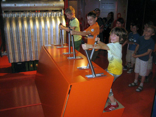 Drei Kinder bedienen große Hebel an einer orangefarbenen Maschine, während andere Kinder in einer Wissenschafts- oder Technologieausstellung zuschauen.