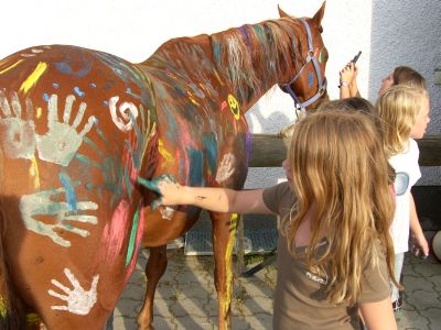 Mehrere Kinder malen bunte Handabdrücke und Muster auf den Körper eines braunen Pferdes, das im Freien neben einer weißen Wand steht.