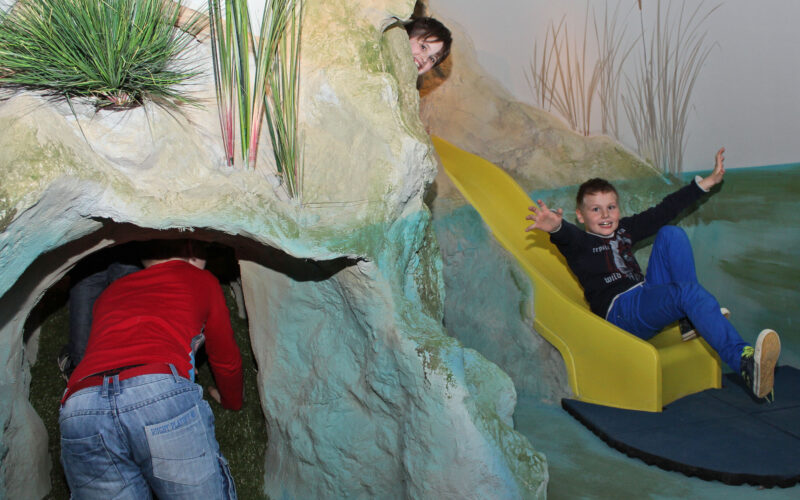 Drei Kinder spielen auf einem Indoor-Spielplatz mit einem Tunnel und einer gelben Rutsche; ein Kind krabbelt in den Tunnel, während zwei andere aus der Rutsche kommen.