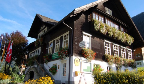 Ein traditionelles Alpengebäude mit dunkler Holzfassade, Blumenkästen an den Fenstern, einer Treppe und Schweizer Flaggen draußen an einem sonnigen Tag.