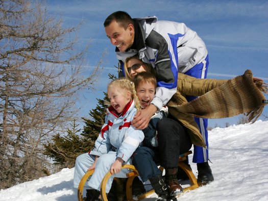 Vier Personen, zwei Erwachsene und zwei Kinder, fahren gemeinsam auf einem Holzschlitten einen verschneiten Hügel hinunter, lächeln und lachen unter blauem Himmel.