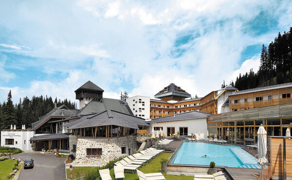 Ein moderner Ferienkomplex in den Bergen mit mehreren Gebäuden, einem Außenpool, Liegestühlen und bewaldeten Hügeln im Hintergrund unter einem teilweise bewölkten Himmel.