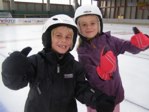 Zwei Kinder mit Helmen und Winterjacken zeigen auf einer Eisbahn den Daumen nach oben.