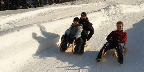 Drei Personen fahren an einem sonnigen Wintertag mit Holzschlitten eine schneebedeckte Bahn hinunter, umgeben von hohen Schneewänden.