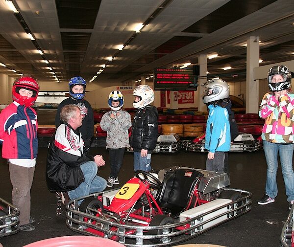 Eine Gruppe von Kindern mit Helmen und ein Mann versammeln sich um Gokarts in einer Indoor-Bahn; ein Kind steht neben einem Gokart mit der Nummer 3.