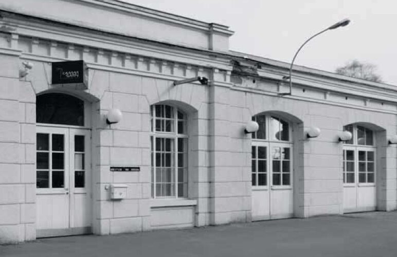 Ein weißes, einstöckiges Gebäude mit gewölbten Fenstern und Türen, Wandleuchten und einer Straßenlaterne davor. Der Himmel scheint bedeckt zu sein.