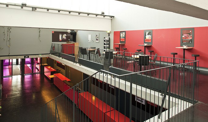 Modernes Zwischengeschoss im Innenbereich mit roten und grauen Wänden, schwarzem Geländer, hohen Tischen mit Hockern und einer Treppe, die in eine untere Etage führt. An den Wänden sind Plakate angebracht.