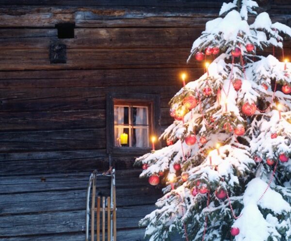 Ein schneebedeckter, mit roten Ornamenten und Kerzen geschmückter Weihnachtsbaum steht vor einer rustikalen Holzhütte mit einem beleuchteten Fenster und einem Schlitten in der Nähe.
