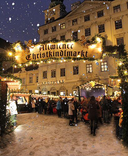 Menschenmengen versammeln sich unter dem beleuchteten Schild "Altwiener Christkindlmarkt" auf einem verschneiten Weihnachtsmarkt im Freien vor einem historischen Gebäude in der Abenddämmerung.