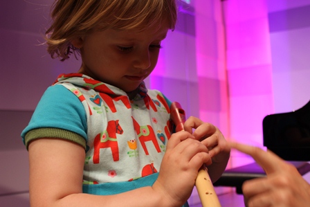 Ein kleines Kind in einem bunten Hemd spielt eine Holzblockflöte, während eine Erwachsenenhand auf das Instrument zeigt. Der Hintergrund ist mit rosa und lila Lichtern beleuchtet.