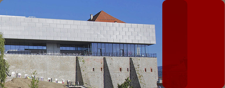 Ein modernes Gebäude mit einer Fassade aus Glas und Metall thront auf einer alten Steinmauer unter einem klaren blauen Himmel. Zwei große rote Rechtecke überlagern die rechte Seite des Bildes.