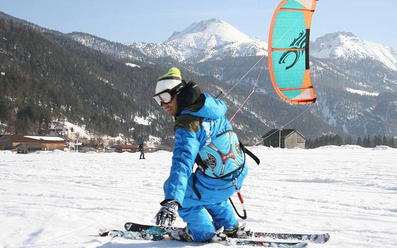 Eine Person in blauer Winterkleidung fährt auf Skiern durch eine verschneite Landschaft mit Bergen und Gebäuden im Hintergrund.