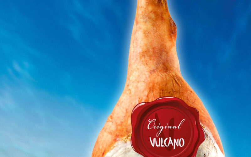Ein ganzer Rohschinken mit einem roten "Original Vulcano"-Etikett hängt vor einem blauen Himmel und einem grünen Landschaftshintergrund.