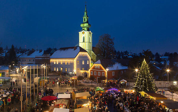 Eine nächtliche Szene eines Weihnachtsmarktes auf einem Stadtplatz mit einer Menschenmenge, geschmückten Ständen, einem beleuchteten Weihnachtsbaum und einer Kirche mit beleuchtetem Uhrenturm im Hintergrund.
