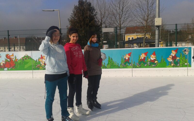 Drei Personen in Winterkleidung stehen auf einer Eislaufbahn im Freien mit einem bunten Gnom-Wandbild auf der Absperrung hinter ihnen.