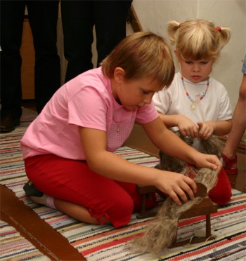 Zwei kleine Kinder knien auf einem gestreiften Teppich und benutzen ein traditionelles Werkzeug, um Rohwolle zu kämmen. Andere Menschen stehen im Hintergrund.