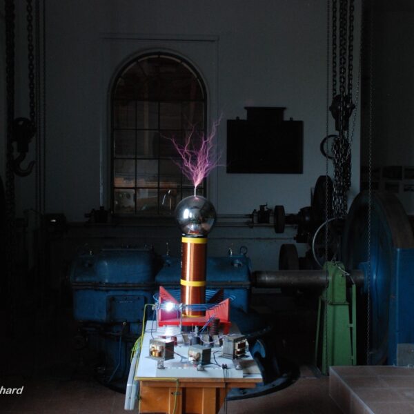 Eine Teslaspule, die violette elektrische Entladungen in einem schwach beleuchteten Industrieraum mit Maschinen und einem großen Fenster im Hintergrund abgibt.