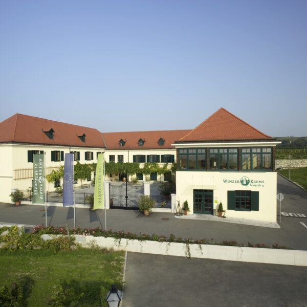 Ein moderner Gebäudekomplex mit roten Ziegeldächern, cremefarbenen Wänden und großen Fenstern, umgeben von einem Parkplatz und Grünflächen unter einem klaren Himmel.
