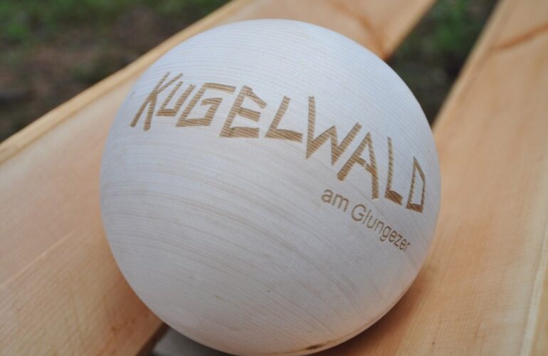Eine glatte Holzkugel mit dem eingravierten Schriftzug "KUGELWALD am Glungezer", die auf einer hellen Holzfläche ruht.