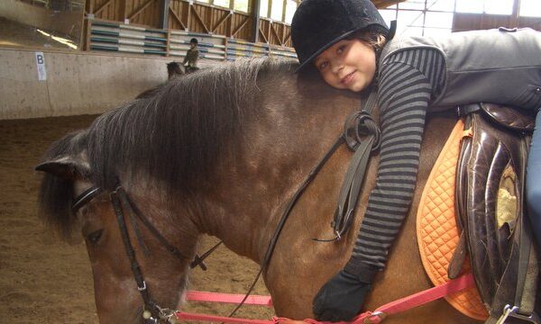 Ein Kind, das einen Helm und Handschuhe trägt, liegt auf dem Rücken eines braunen Pferdes mit orangefarbener Schabracke in einer Reithalle.