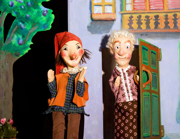 Zwei Puppen, die ein Mädchen und eine ältere Frau darstellen, stehen vor einem bemalten Haus mit einem Baum und Blumen im Hintergrund.