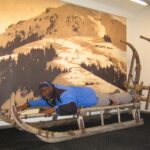 Ein Mann liegt mit dem Gesicht nach unten auf einem großen Holzschlitten im Haus und posiert, als würde er darauf fahren.