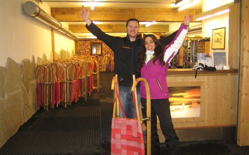 Zwei Personen stehen in einem Haus mit erhobenen Armen und lächelnd neben einem Holzschlitten. Im Hintergrund sind Reihen ähnlicher Schlitten aufgereiht. Die Umgebung scheint ein Verleih oder eine Ausrüstungsfirma zu sein.