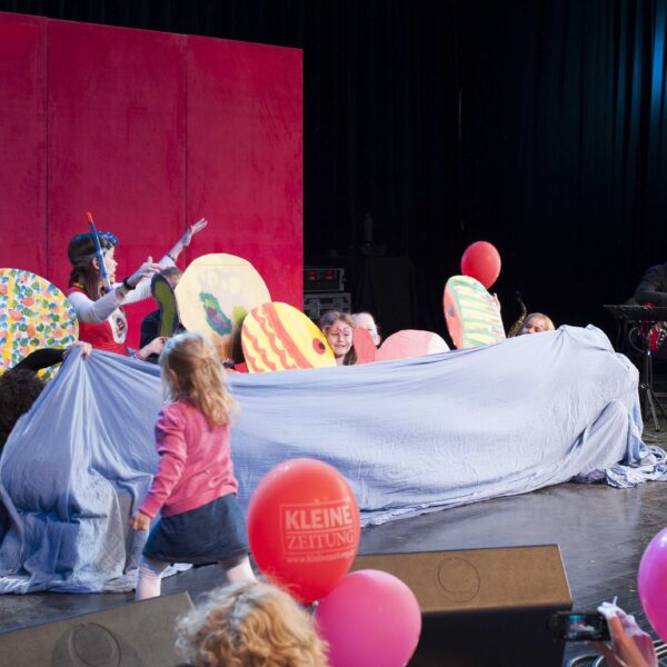 Kinder und Erwachsene treten auf der Bühne mit bunten Fischrequisiten und einem blauen Tuch auf, während ein Musiker nebenbei Gitarre spielt; Zuschauer mit Luftballons schauen zu.