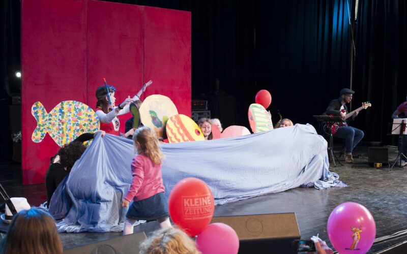 Kinder und Erwachsene treten auf der Bühne mit bunten Fischrequisiten und einem blauen Tuch auf, während ein Musiker nebenbei Gitarre spielt; Zuschauer mit Luftballons schauen zu.