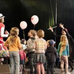 Eine Gruppe von Kindern versammelt sich auf der Bühne, während zwei Erwachsene in Kostümen, einer mit einem Umhang und einem Zauberstab, mit ihnen interagieren. Einige Kinder halten Luftballons; der Hintergrund ist durch die Bühnenbeleuchtung dunkel.