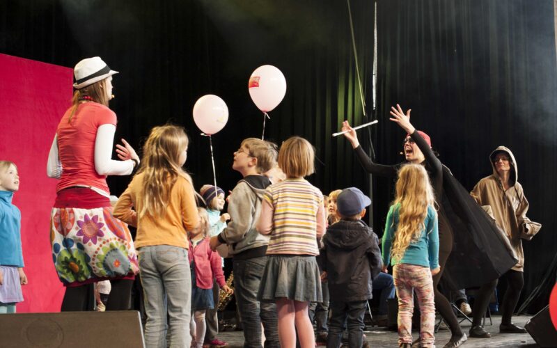 Eine Gruppe von Kindern versammelt sich auf der Bühne, während zwei Erwachsene in Kostümen, einer mit einem Umhang und einem Zauberstab, mit ihnen interagieren. Einige Kinder halten Luftballons; der Hintergrund ist durch die Bühnenbeleuchtung dunkel.