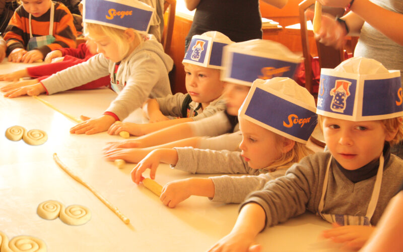Mehrere kleine Kinder mit Papierhüten und Schürzen formen während einer Backaktion auf einem Tisch Teig zu Spiralen.