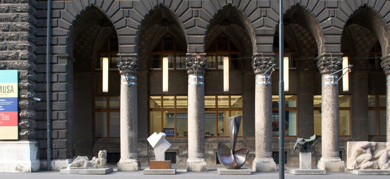 Eine Reihe von Steinsäulen steht vor einer bogenförmigen Gebäudefassade mit mehreren modernen Skulpturen und einem Schild mit der Aufschrift "MUSA" auf der linken Seite.