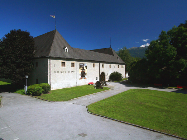Ein weißes Steingebäude mit dunklen Dächern und der Aufschrift "Museum Zeughaus" steht umgeben von grünen Rasenflächen, Bäumen und einem Gehweg unter einem klaren blauen Himmel.