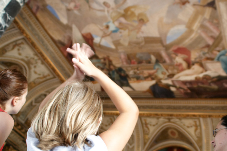 Zwei Personen, die von hinten gesehen werden, heben ihre Hände in Richtung einer bemalten Decke in einem kunstvoll verzierten Gebäude.