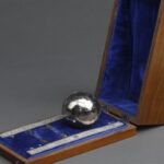 Eine silberne Fußballtrophäe in einer mit blauem Samt ausgekleideten Holzkiste, die offen auf einem grauen Hintergrund ausgestellt ist.