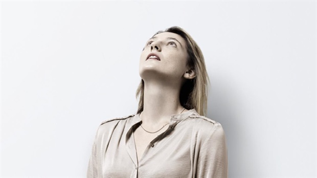 Frau mit hellbraunem Haar und beiger Bluse blickt nach oben und steht vor einem schlichten weißen Hintergrund.