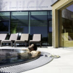 Ein Mann entspannt sich in einem runden Außenpool neben mehreren Liegestühlen und einem modernen Gebäude mit großen Glasfenstern.