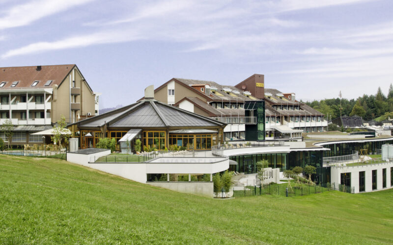 Ein moderner Hotelkomplex mit mehreren miteinander verbundenen Gebäuden und großen Fenstern, der auf einem grasbewachsenen Hügel unter einem teilweise bewölkten Himmel liegt.
