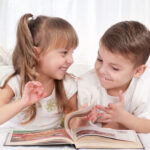 Zwei kleine Kinder liegen nebeneinander auf einem Bett, lächeln und schauen sich an, während sie gemeinsam in einem Buch blättern.