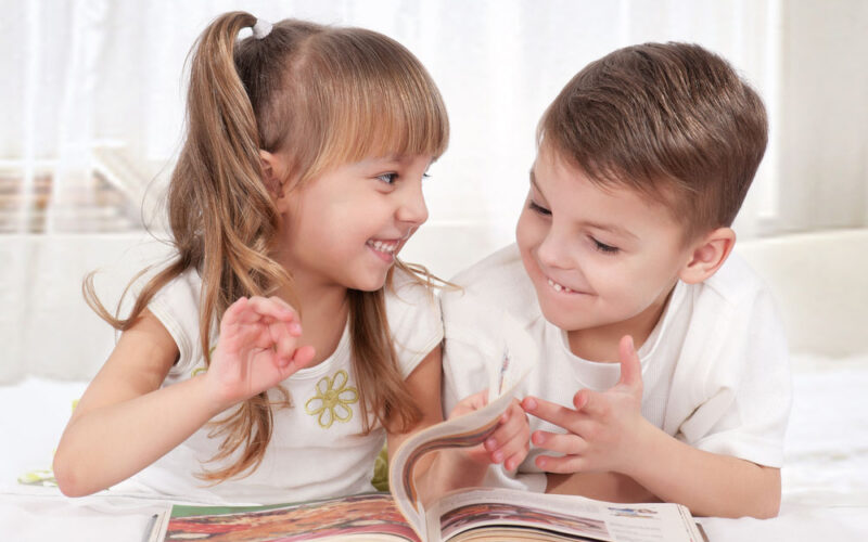 Zwei kleine Kinder liegen nebeneinander auf einem Bett, lächeln und schauen sich an, während sie gemeinsam in einem Buch blättern.