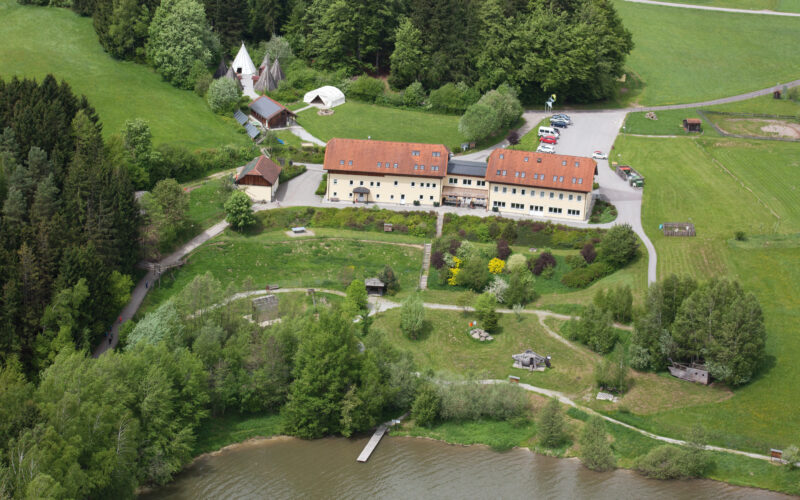 Luftaufnahme eines großen gelben Gebäudes mit rotem Dach, umgeben von Bäumen, offenen Feldern, geparkten Autos, Zelten und einem Steg, der in einen See hineinragt.
