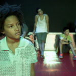 Eine junge Person mit lockigem Haar steht im Vordergrund, während vier weitere Personen im Hintergrund auf einem roten Boden sitzen oder an einer Wand lehnen und nachdenklich wirken.