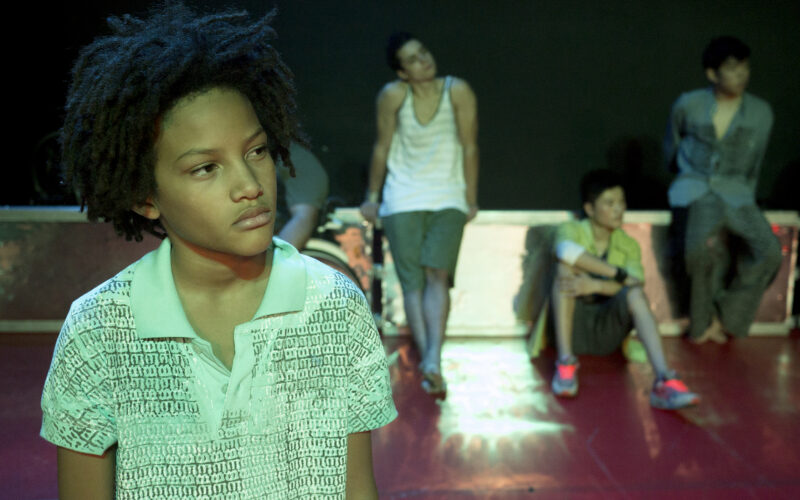 Eine junge Person mit lockigem Haar steht im Vordergrund, während vier weitere Personen im Hintergrund auf einem roten Boden sitzen oder an einer Wand lehnen und nachdenklich wirken.