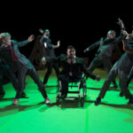 Eine Gruppe von Darstellern in Anzügen, darunter eine Person im Rollstuhl, posiert dramatisch auf einer grün beleuchteten Bühne.