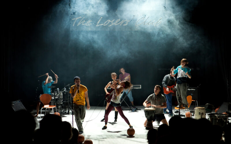 Eine Band spielt energisch auf der Bühne unter dramatischer Beleuchtung, mit verschiedenen Instrumenten und einem Schild mit der Aufschrift "The Lovers Club" im Hintergrund.