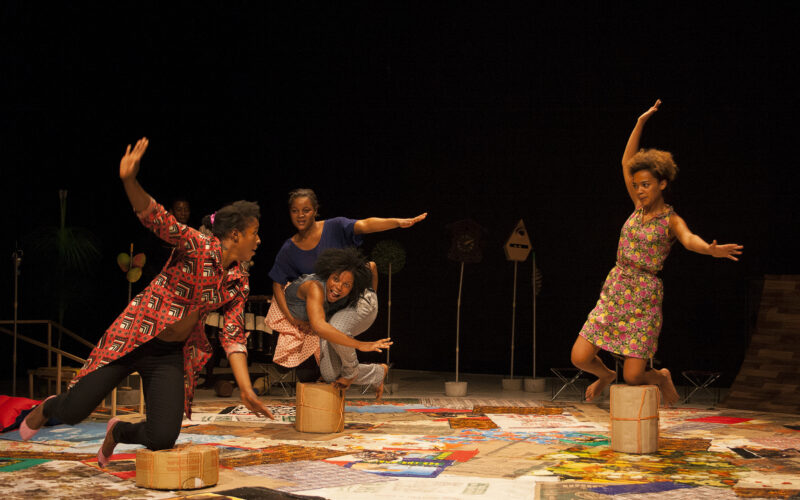 Vier Personen treten energisch auf der Bühne auf, balancieren auf kleinen Hockern, tragen bunte Outfits und einen gemusterten Boden vor einem dunklen Hintergrund.