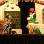 Eine Puppenspielszene mit einer Hundepuppe, die einer menschlichen Puppe in bunten Kleidern und einem roten Hut gegenübersteht, in einer Küche mit Tisch, Tellern und Herd.