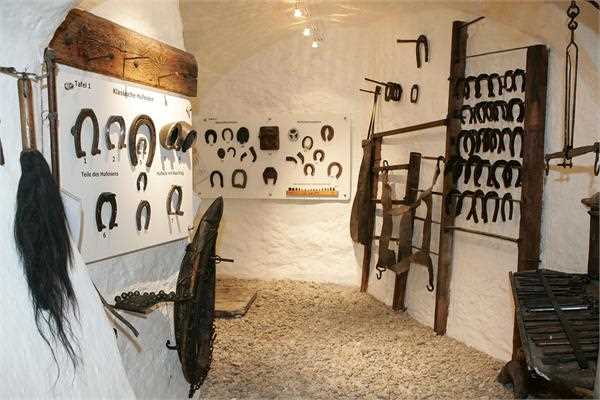 Ausstellungsraum des Museums mit verschiedenen alten Hufeisen, Schmiedewerkzeugen und Ausrüstungsgegenständen, die an weißen Wänden und in Holzregalen ausgestellt sind, wobei der Boden mit Kies bedeckt ist.