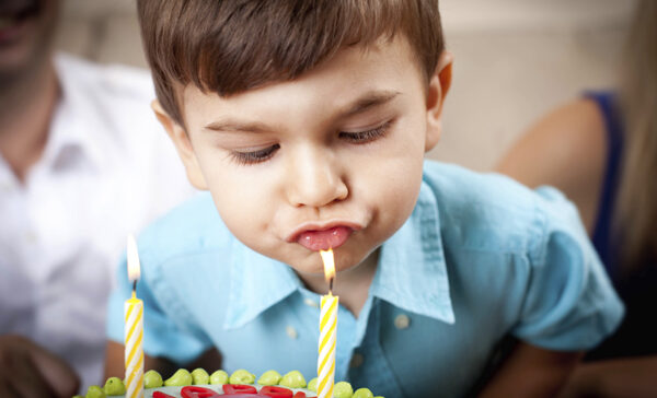 Ein kleiner Junge in einem blauen Hemd bläst zwei brennende Kerzen auf einer Geburtstagstorte aus.